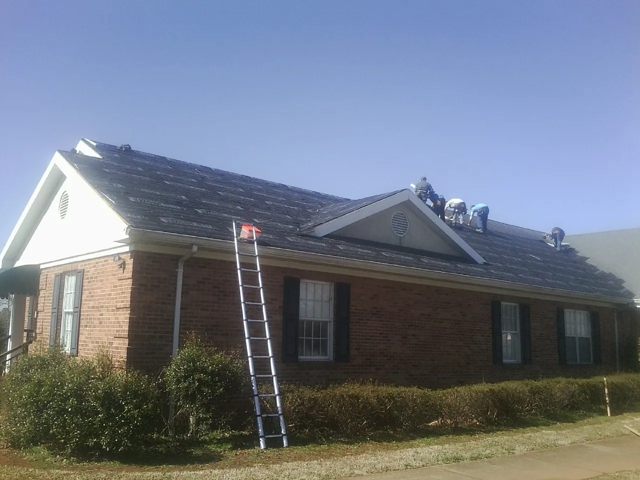 Crew tearing shingles off a brick ranch roof with a ladder set — Emergency Roof Repair work by our crew in Omega