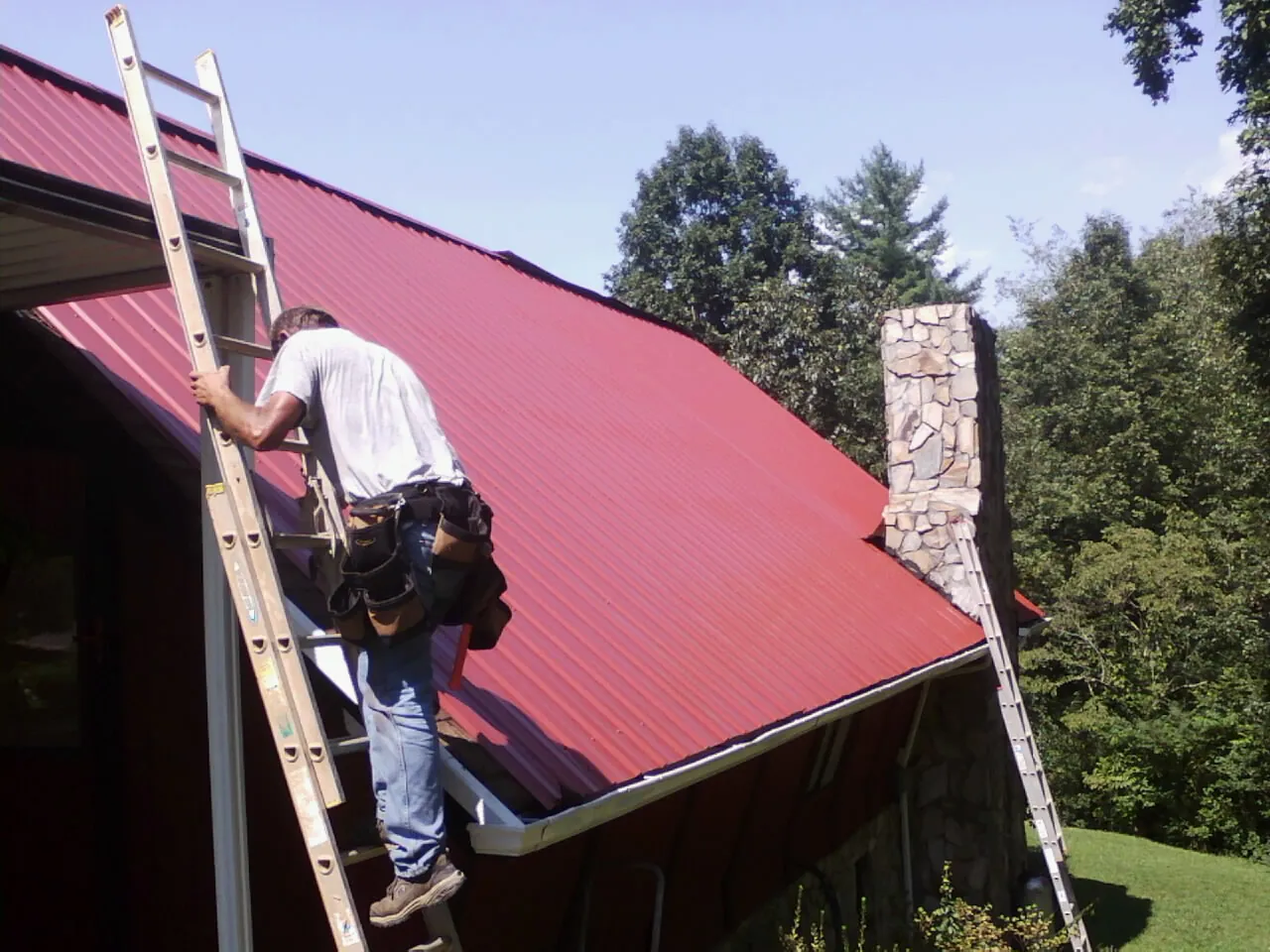 Roofer on a ladder beside a red metal roof and stone chimney — Soffit & Fascia Repair by Roofing Brothers in Omega
